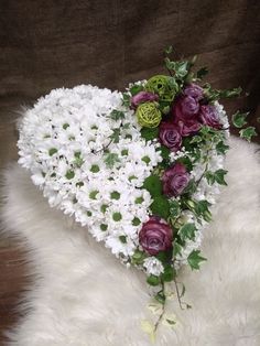 a heart - shaped arrangement of white and purple flowers on a sheepskin rug in front of a brown background