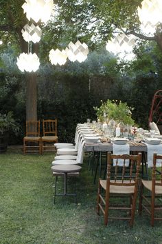 an outdoor dining table with chairs and lights hanging from the ceiling over it, surrounded by greenery