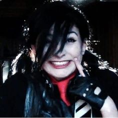 a woman wearing black leather gloves and a red shirt smiles at the camera with her hand on her face