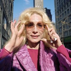 a woman with blonde hair and glasses is holding her hands up to her face while standing in front of tall buildings