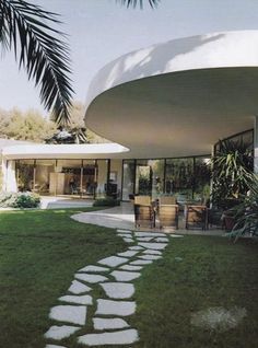 the house is surrounded by lush green grass and palm trees, with stepping stones leading up to the front door