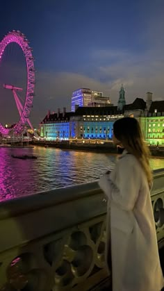 a woman standing on a bridge looking at the water with a ferris wheel in the background