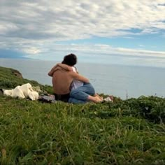 a man and woman sitting on top of a lush green hillside next to the ocean