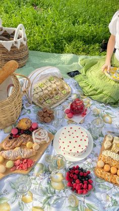 a woman sitting at a picnic table filled with food and desserts next to a basket full of fruit