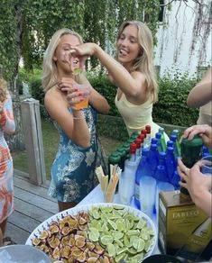 three women standing around a table full of food and drinks, one holding a drink in her hand