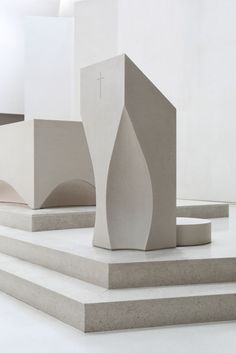 an abstract sculpture sits on concrete steps in front of a white wall and cross at the top