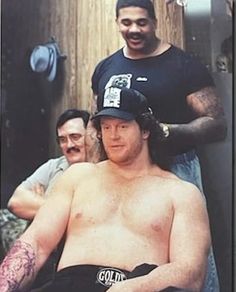 a man with no shirt on getting his hair cut by another man wearing a hat