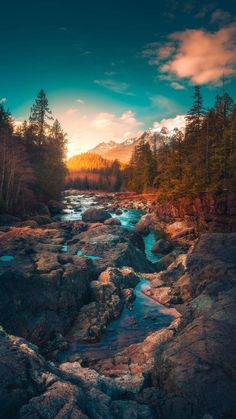 a river running through a forest filled with rocks under a cloudy blue sky at sunset