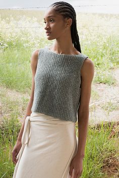 a woman standing in the grass wearing a gray sweater and white skirt with braids