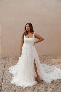 a woman standing in front of a wall wearing a white wedding dress with high slit