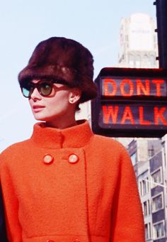 a woman in an orange coat and sunglasses standing next to a don't walk sign