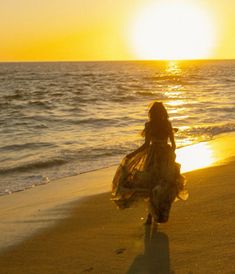 a woman walking on the beach at sunset with her dress blowing in the wind,