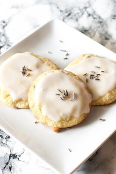 three cookies with icing and lavender on a white plate sitting on a marble table