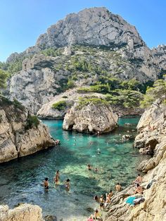 many people are swimming in the clear blue water near some rocks and trees, with mountains in the background