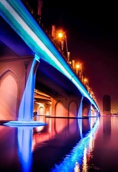 a bridge that is lit up at night with lights reflecting on the water and buildings in the background