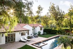 an outdoor pool surrounded by greenery and trees in front of a white house with red tiled roof