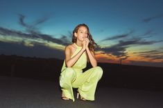 a woman kneeling down on the ground in front of a sky with clouds and sunset behind her