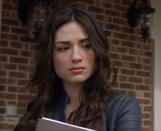 a woman holding a book in front of a brick wall and looking at the camera