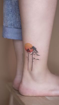 a woman's foot with a small tree tattoo on her left side leg and an orange sunset behind the trees