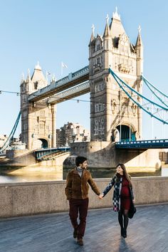 a man and woman holding hands while walking in front of the tower bridge, london