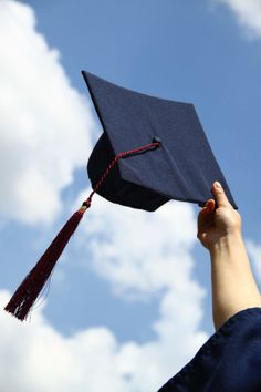 a graduation cap being held up in the air by someone's hand, against a blue sky with white clouds