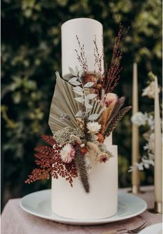 a white wedding cake with dried flowers and feathers on the top, surrounded by candles