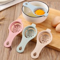 four different colored measuring spoons on a cutting board with eggs in the bowl behind them