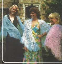 three women standing next to each other in front of a brick building wearing crocheted shawls