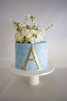 a blue cake with white flowers and a letter on the top is sitting on a plate