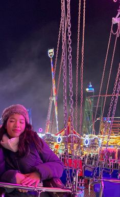 a woman sitting on a bench in front of a ferris wheel at night with lights and carnival rides behind her