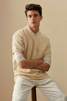 a young man sitting on top of a chair wearing white pants and a beige sweater