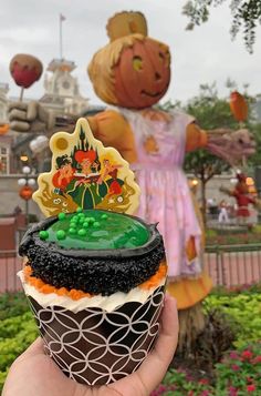 a hand holding up a cupcake in front of a fake scarecrow and pumpkin