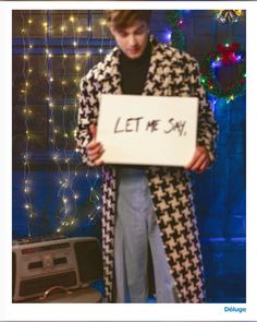 a man holding a sign that says let me say on it in front of a christmas tree