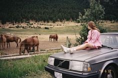 a woman sitting on the hood of a car in front of some horses behind a fence