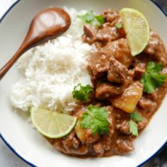 a white plate topped with meat and rice next to a wooden spoon on top of a table