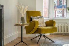 a yellow chair sitting in front of a window next to a table with a vase on it