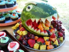 a watermelon shaped like a dinosaur with fruit in it's mouth and cupcakes on the side