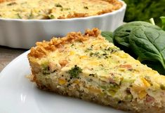 a slice of quiche on a plate next to spinach leaves and a bowl