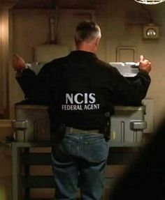 a man standing in front of a counter with his back turned to the camera, wearing a black jacket that reads ncis federal agent