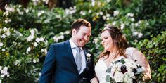 a bride and groom smile as they stand in front of some bushes with white flowers