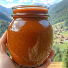 a hand holding a jar filled with sauce in front of a mountain valley and village