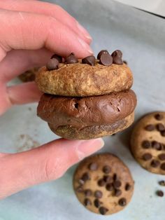 a person holding a chocolate chip cookie doughnut in front of three cookies on a baking sheet