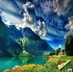 a lake with mountains in the background and green grass on both sides, under a cloudy blue sky