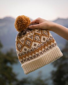 a hand holding onto a brown and white knitted hat with mountains in the background