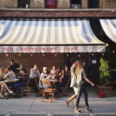 people are sitting at tables in front of a restaurant with awnings over it