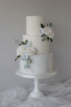 a three tiered cake with white flowers and leaves on the top is sitting on a pedestal