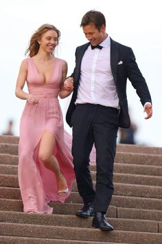 a man and woman are walking down some steps wearing formal wear, one in a pink dress the other in a black tuxedo