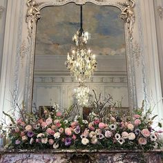 a chandelier hanging from the ceiling above a mantle with flowers and branches in front of it