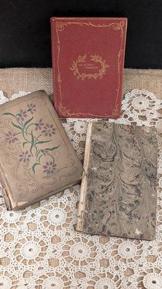 three antique books are sitting on a lace covered tablecloth, one is red and the other is brown