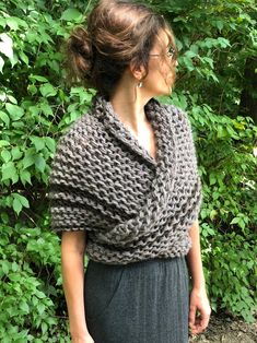 a woman wearing a gray knitted shawl in front of some trees and bushes